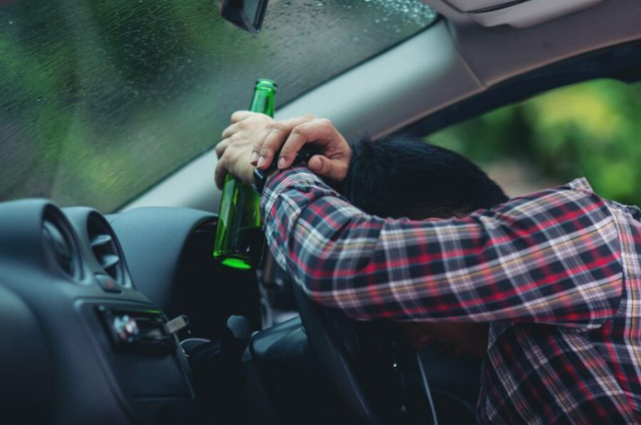 A person laying over a steering wheel with a bottle in his hand. 
